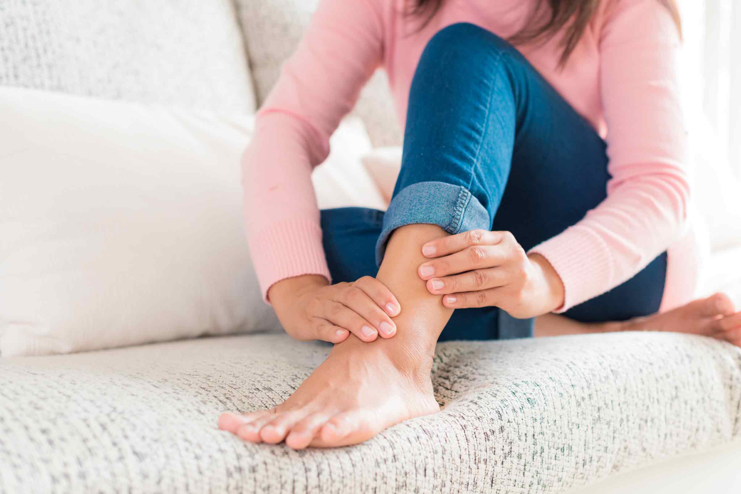 Imagem de uma mulher sentada em um sofá e se segurando o seu tornozelo dolorido